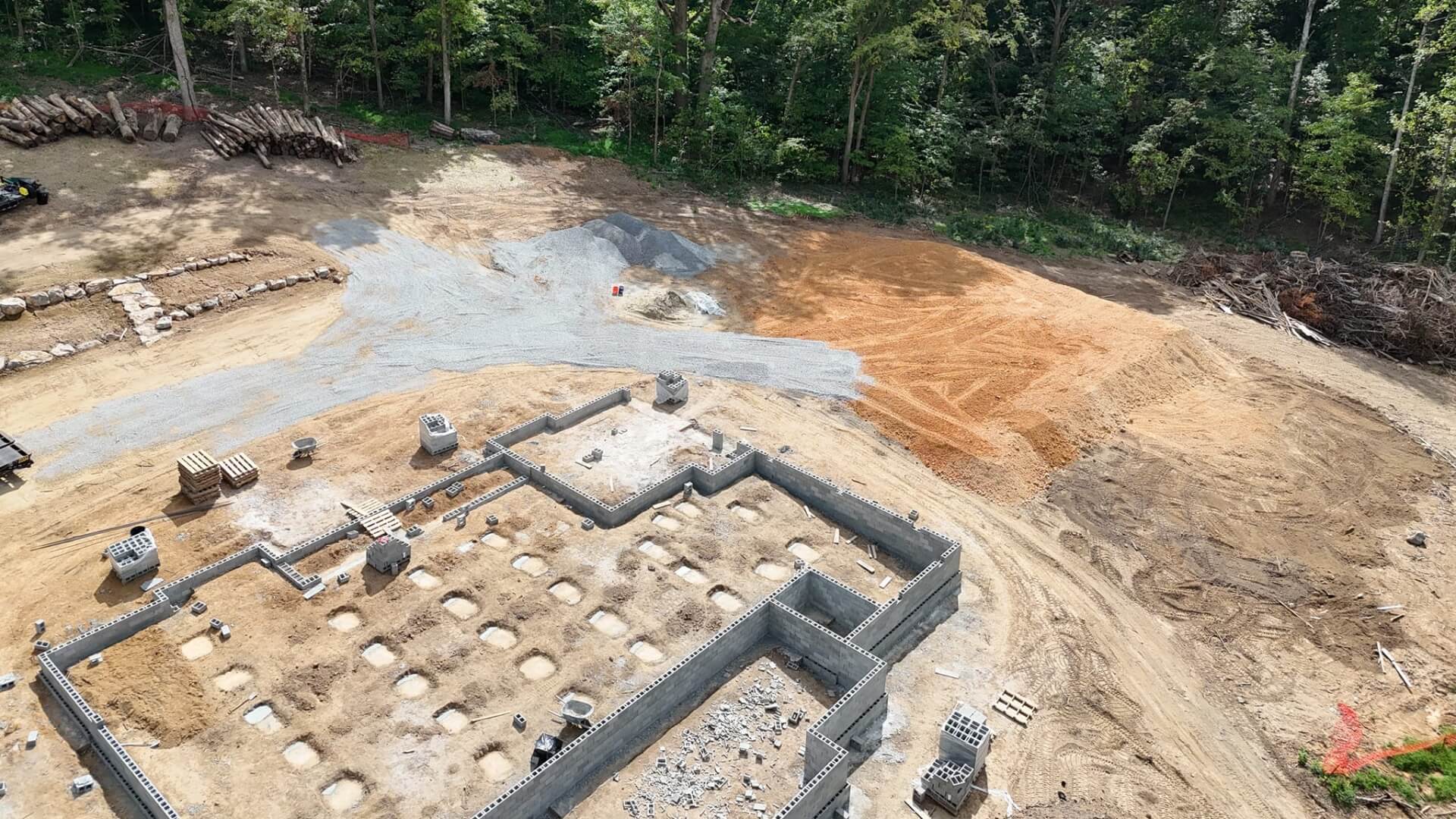 A construction site with a building foundation in progress, surrounded by dirt, gravel, stacked blocks, and forested trees in the background.