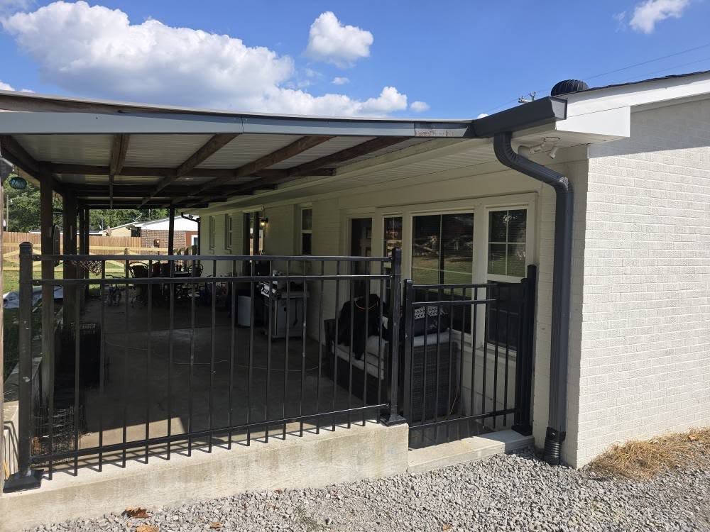 A covered patio attached to a white house, enclosed by a black metal fence. The patio has outdoor furniture and a grill, with a gravel area in front. Blue sky and clouds are visible above.